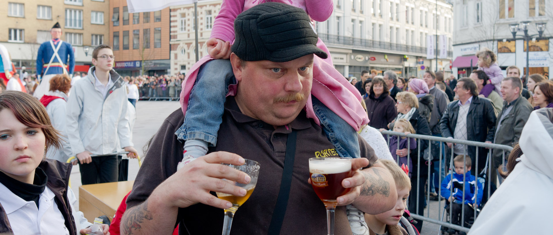Homme avec un enfant sur les épaules et deux verres de bière à la main, au carnaval de Tourcoing, place Victor Hassebroucq.
